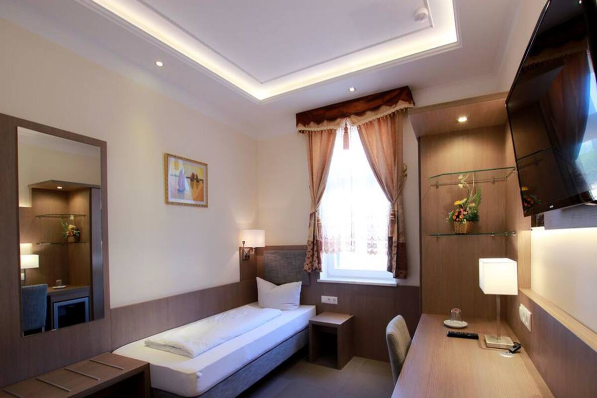 A hotel room with a bed, bedside table, lamp, TV, and a small desk area.