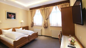 A hotel room with a bed, a TV mounted on the wall, a window with curtains, a radiator, and a framed picture on the wall. - Hotel Europäischer Hof (Elsterwerda)