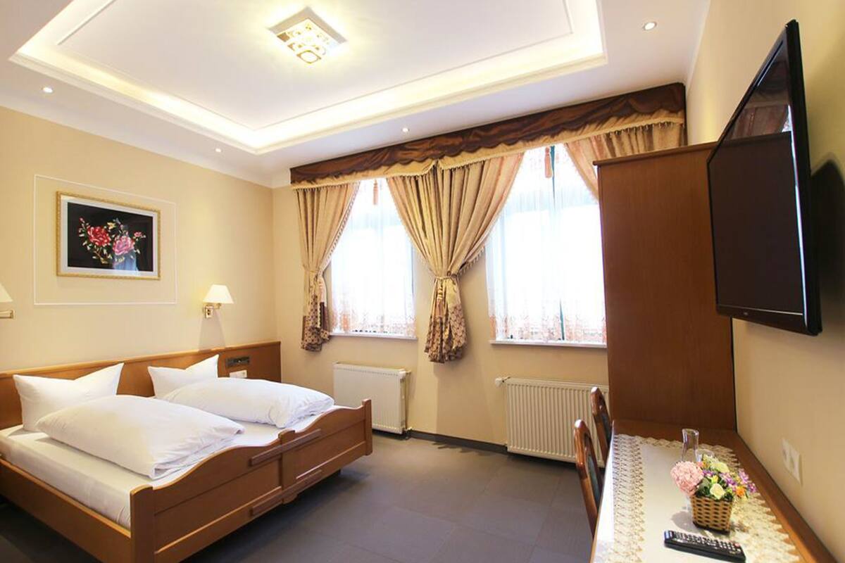 A hotel room with a bed, a television, a window with curtains, a radiator, and a framed picture on the wall.