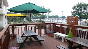 Een houten terras met picknicktafels en een groene parasol, met uitzicht op een parkeerplaats en gebouwen.