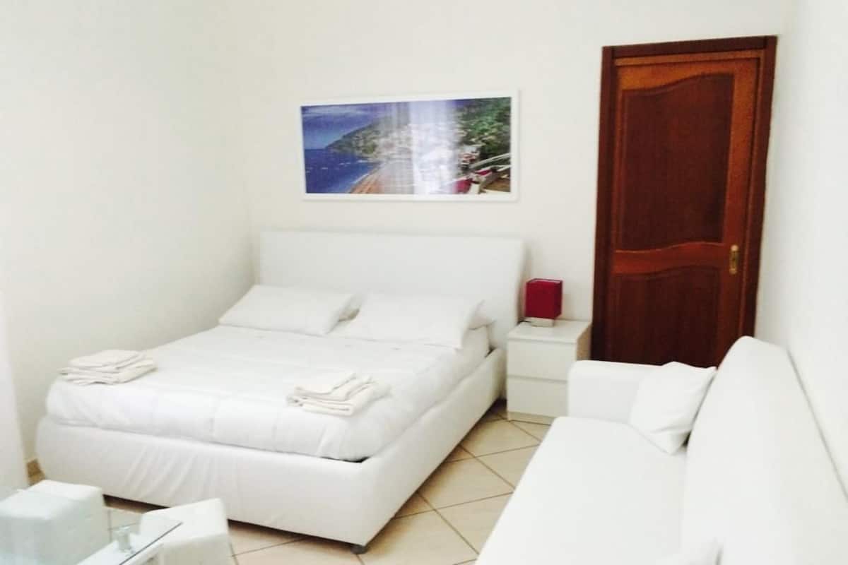 A bedroom with a bed, a bedside table, a nightstand, a white sofa, and a wooden door.