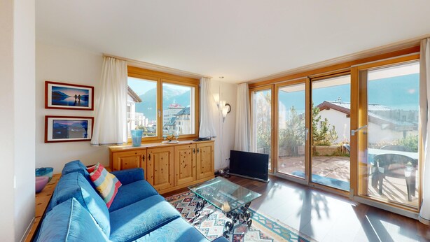 Amplia sala de estar con un sofá azul, armarios de madera, una mesa de centro de vidrio y grandes puertas correderas que dan a un balcón con vistas al mar y edificios.