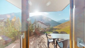 Un balcón con mesa y sillas, con vistas a un paisaje montañoso que incluye una casa y árboles.
