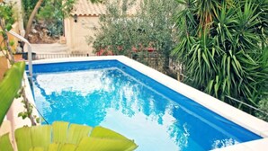 En rektangulær swimmingpool med krystalklart blåt vand, omgivet af frodig vegetation og en fliseklædt bygning i baggrunden.