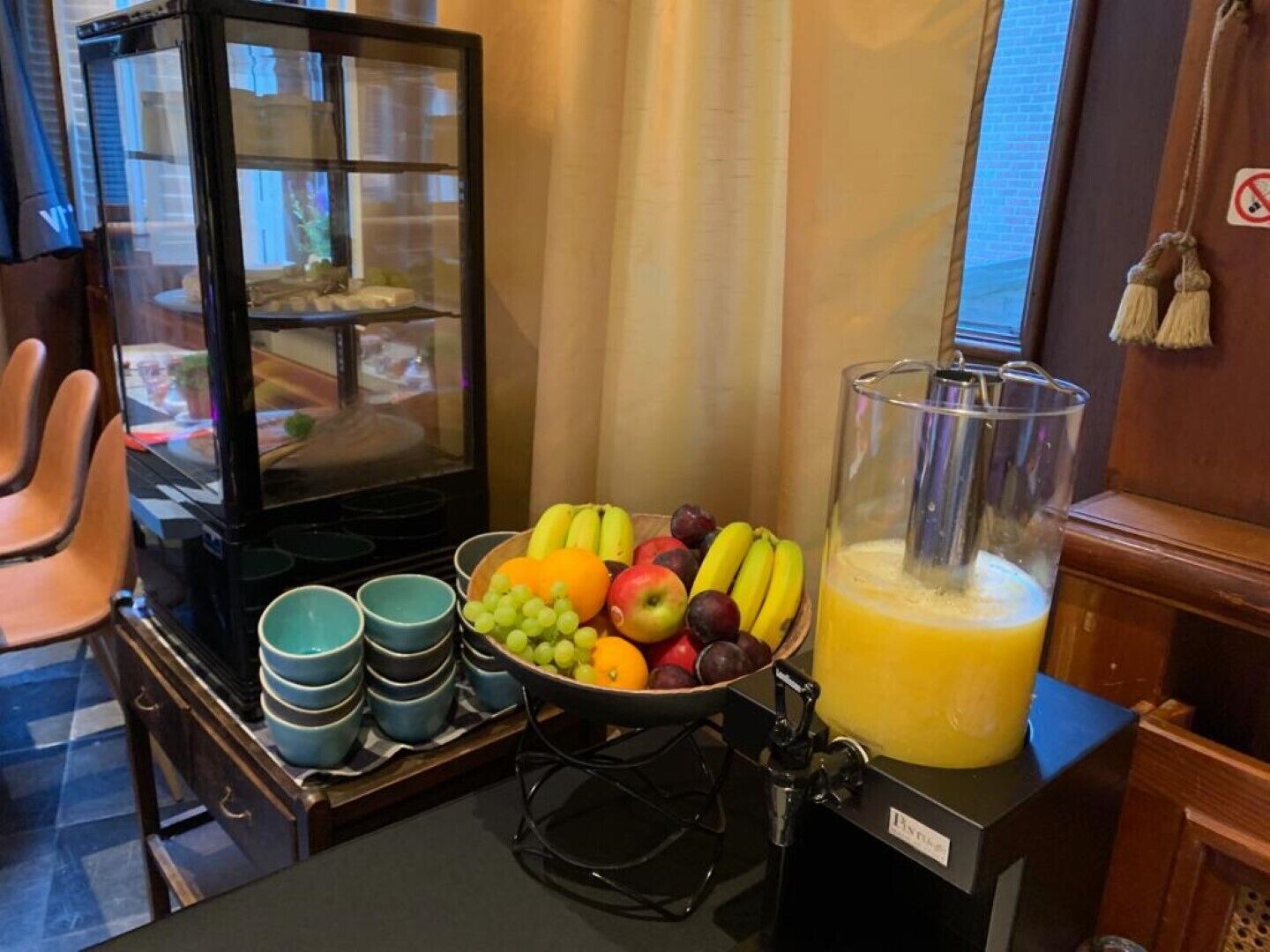 A breakfast setup with a fruit bowl, a blender with orange juice, and stacked blue cups on a table.