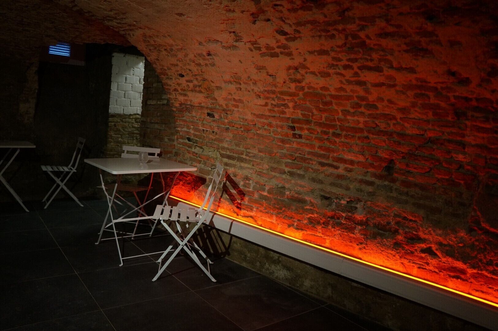 Ein schwach beleuchteter Raum mit Backsteinwänden, weißen Faltstühlen und einem Tisch.