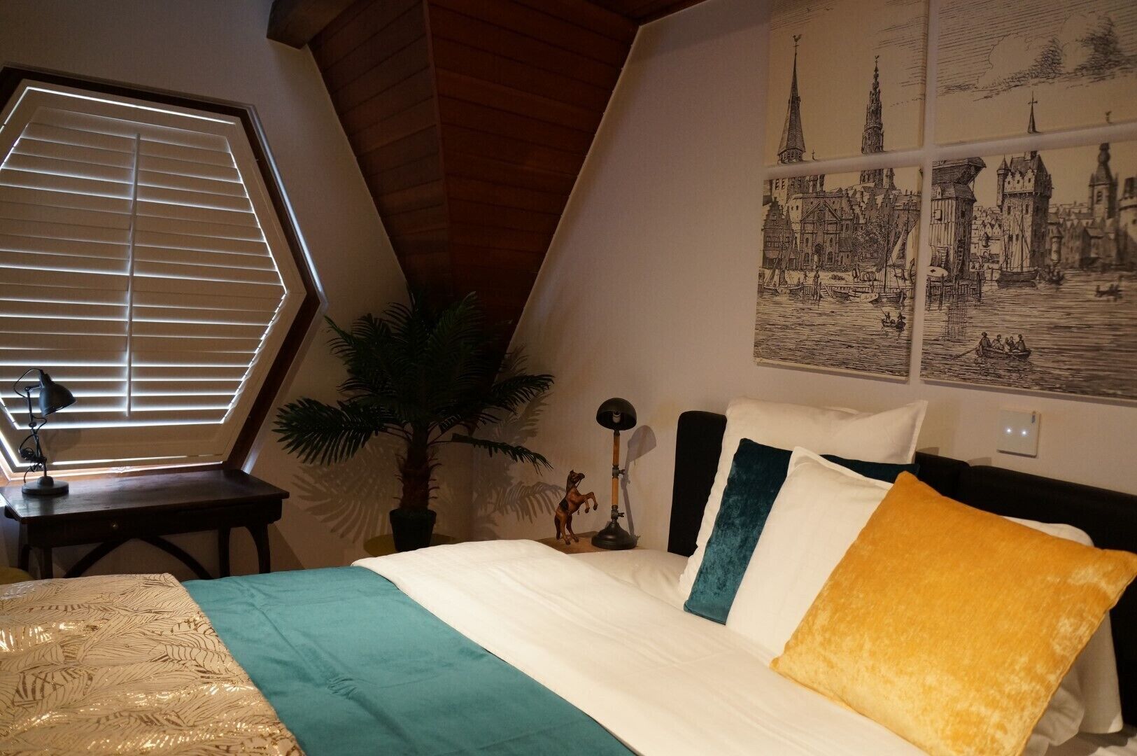 Ein Schlafzimmer mit Bett, Schreibtisch, Lampe, Pflanze und einer Wand mit Kunstwerken.
