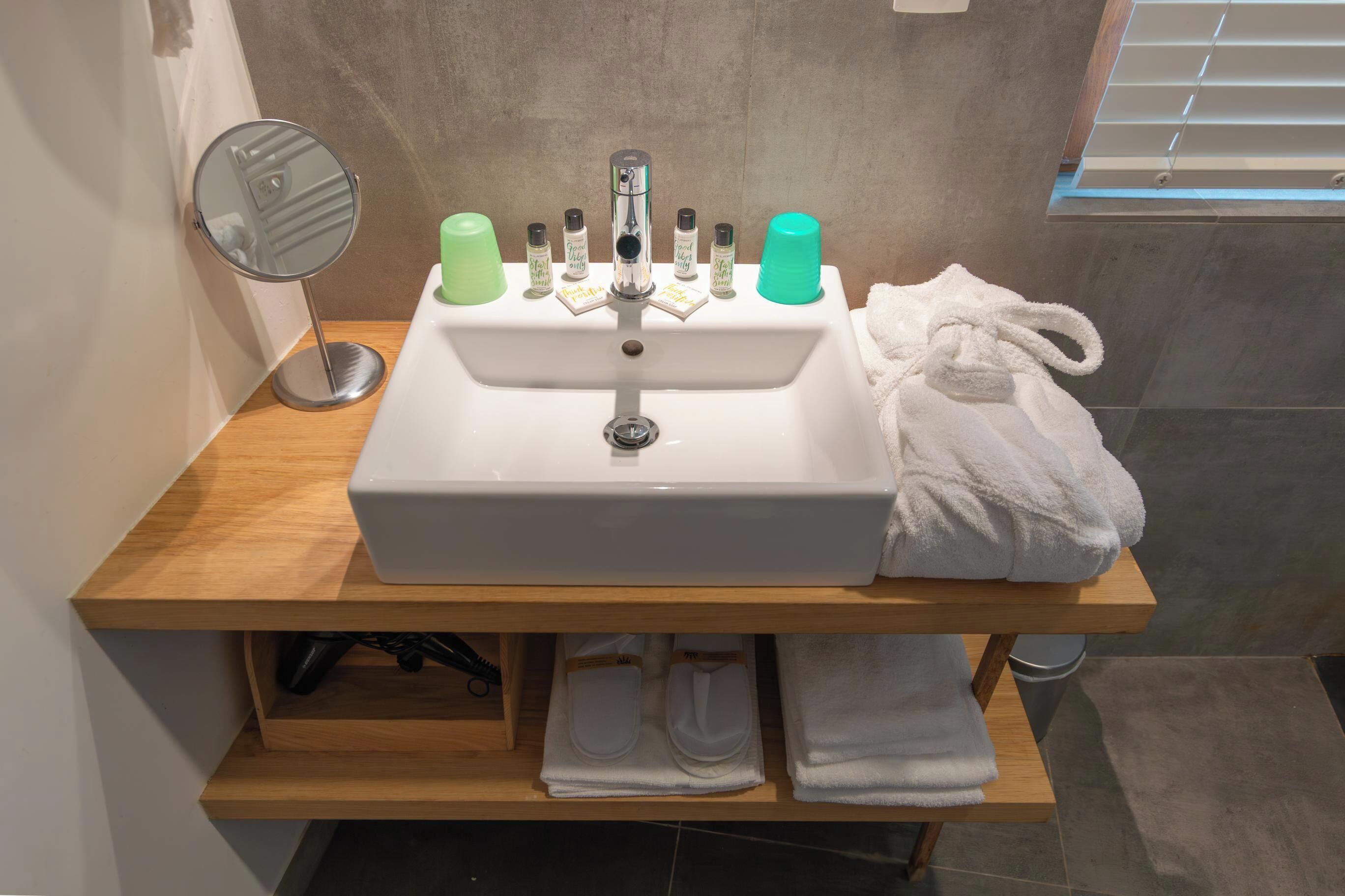 A bathroom sink with toiletries, a mirror, and towels on a wooden shelf.