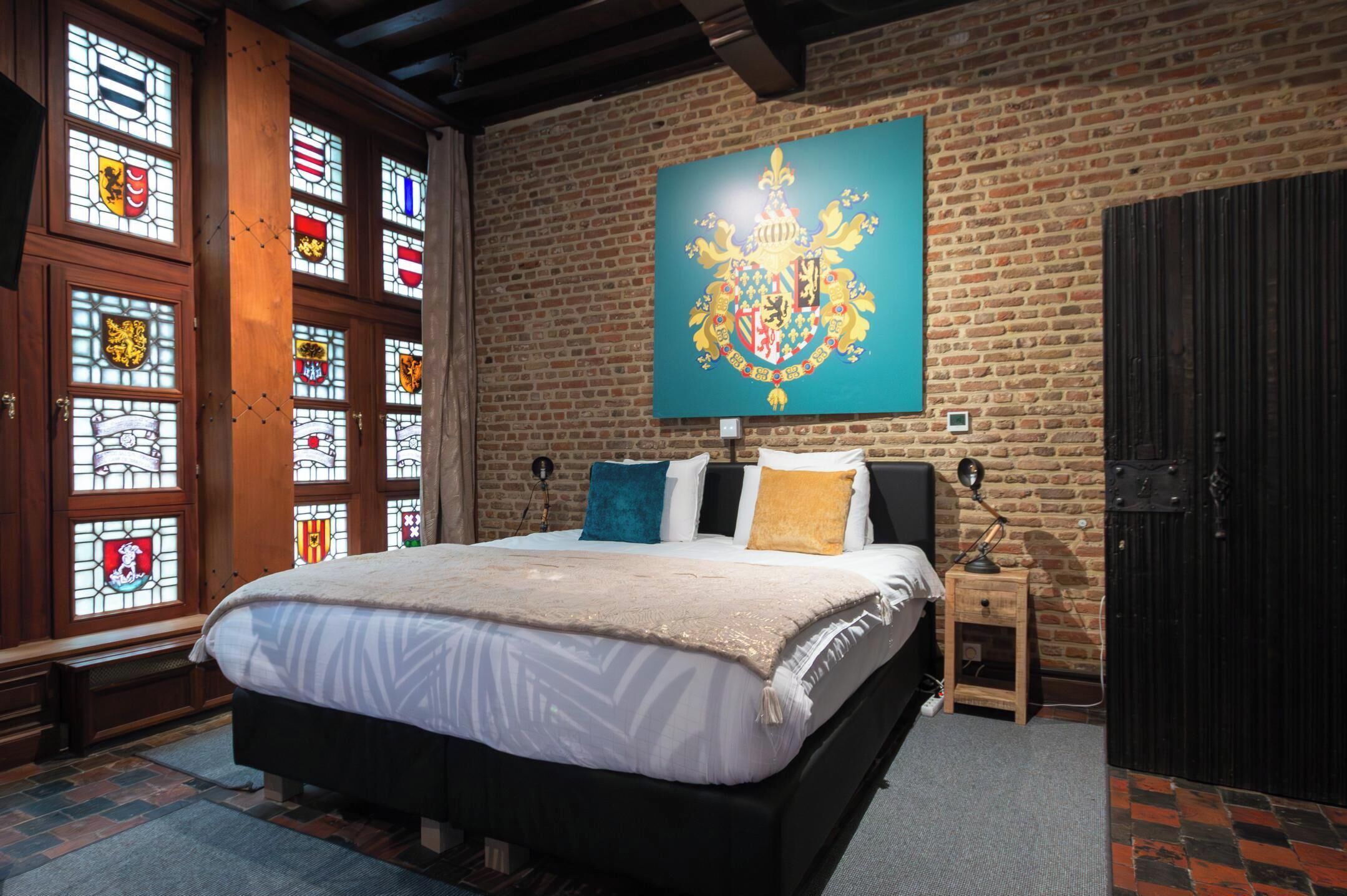 Un dormitorio con una cama grande, un armario de madera, una mesita de noche y un ventanal con vidrieras de motivos heráldicos.