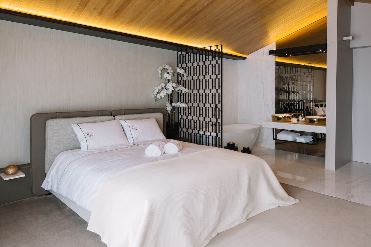 A modern hotel room with a large bed, a wooden ceiling, a decorative partition, and a bathroom with a mirror and sink.