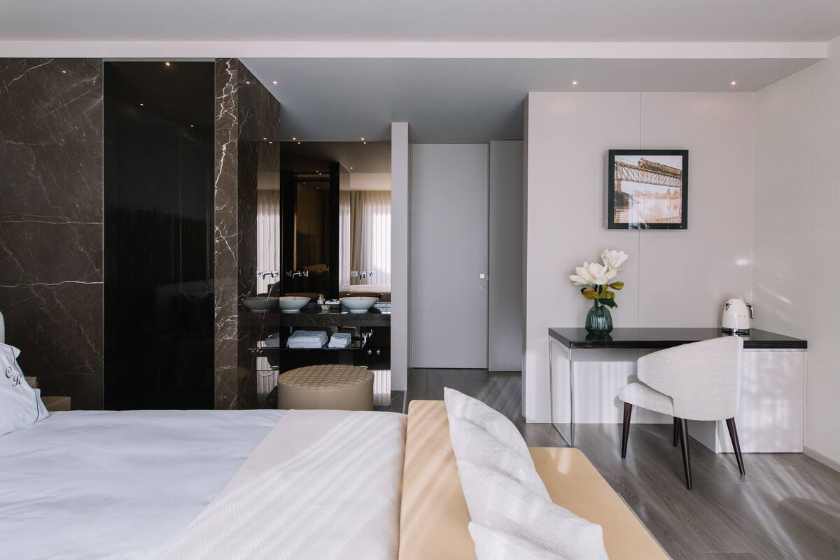 A modern hotel room with a large bed, a sitting area with a chair, a small desk, and a bathroom visible through an open door.