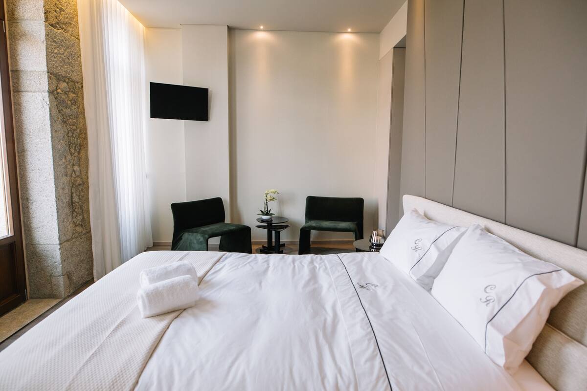 A hotel room with a large bed, two chairs, a small table, and a flat-screen TV on the wall.