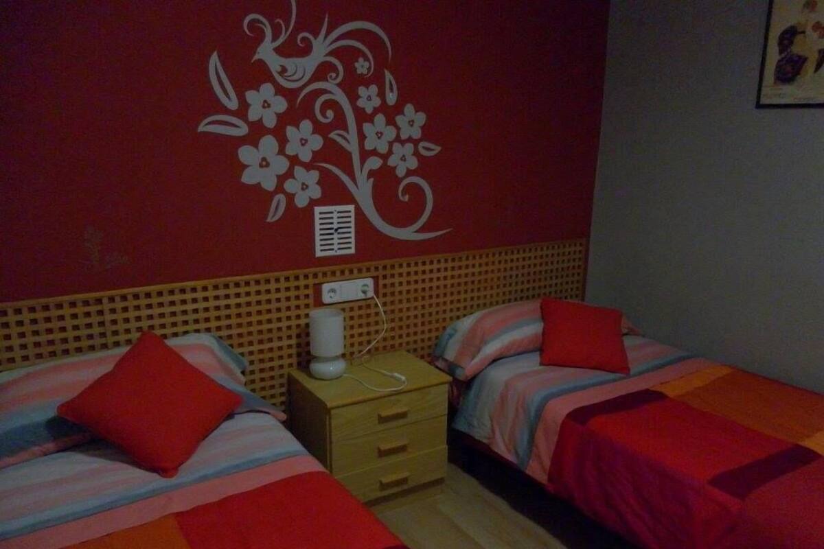 Ett rum med två sängar, var och en med en röd kudde och randigt sänglinne. Mellan sängarna står ett nattduksbord med en lampa och ett eluttag.