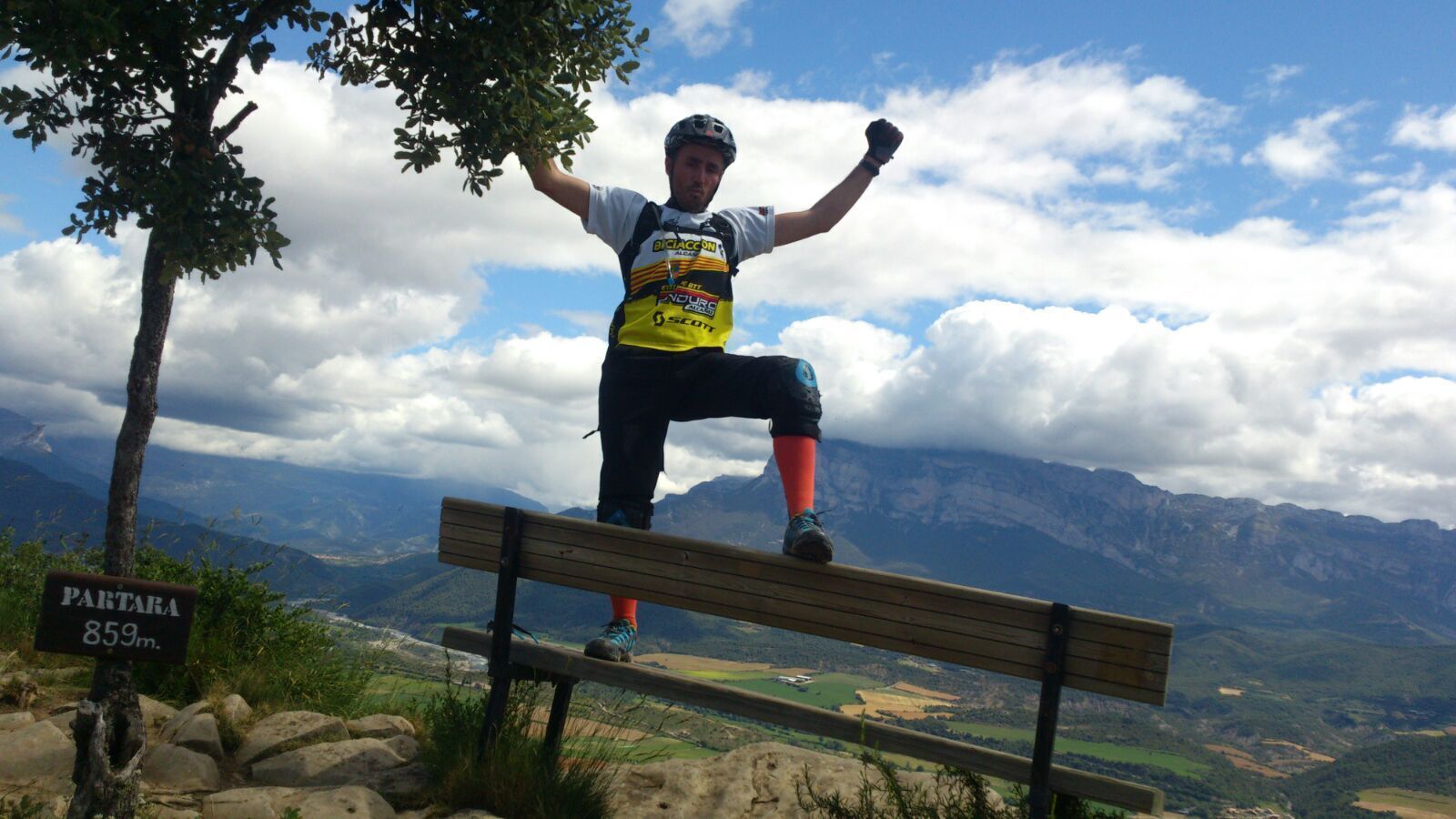 Una persona de pie en un banco de madera, con un paisaje montañoso al fondo.