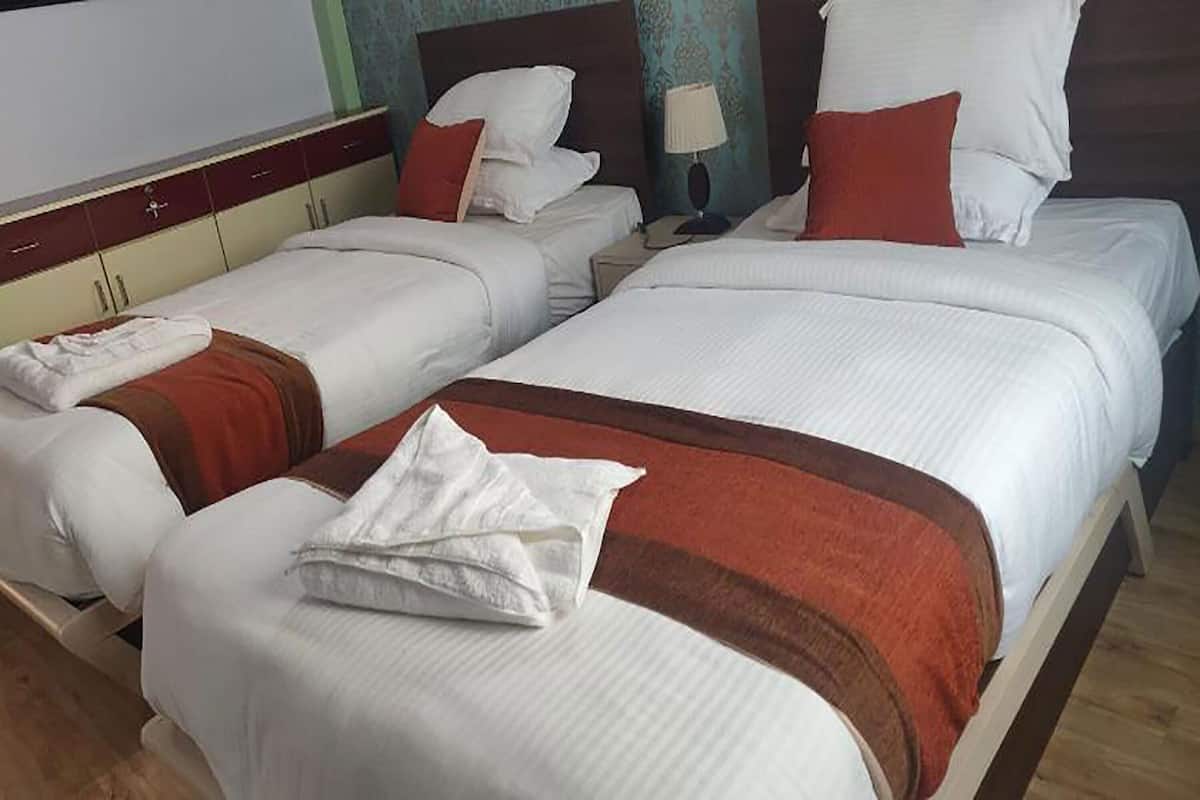 A hotel room with three beds, each with white bedding and red accents. There is a wooden headboard, a bedside table with a lamp, and a wardrobe with a mirror.