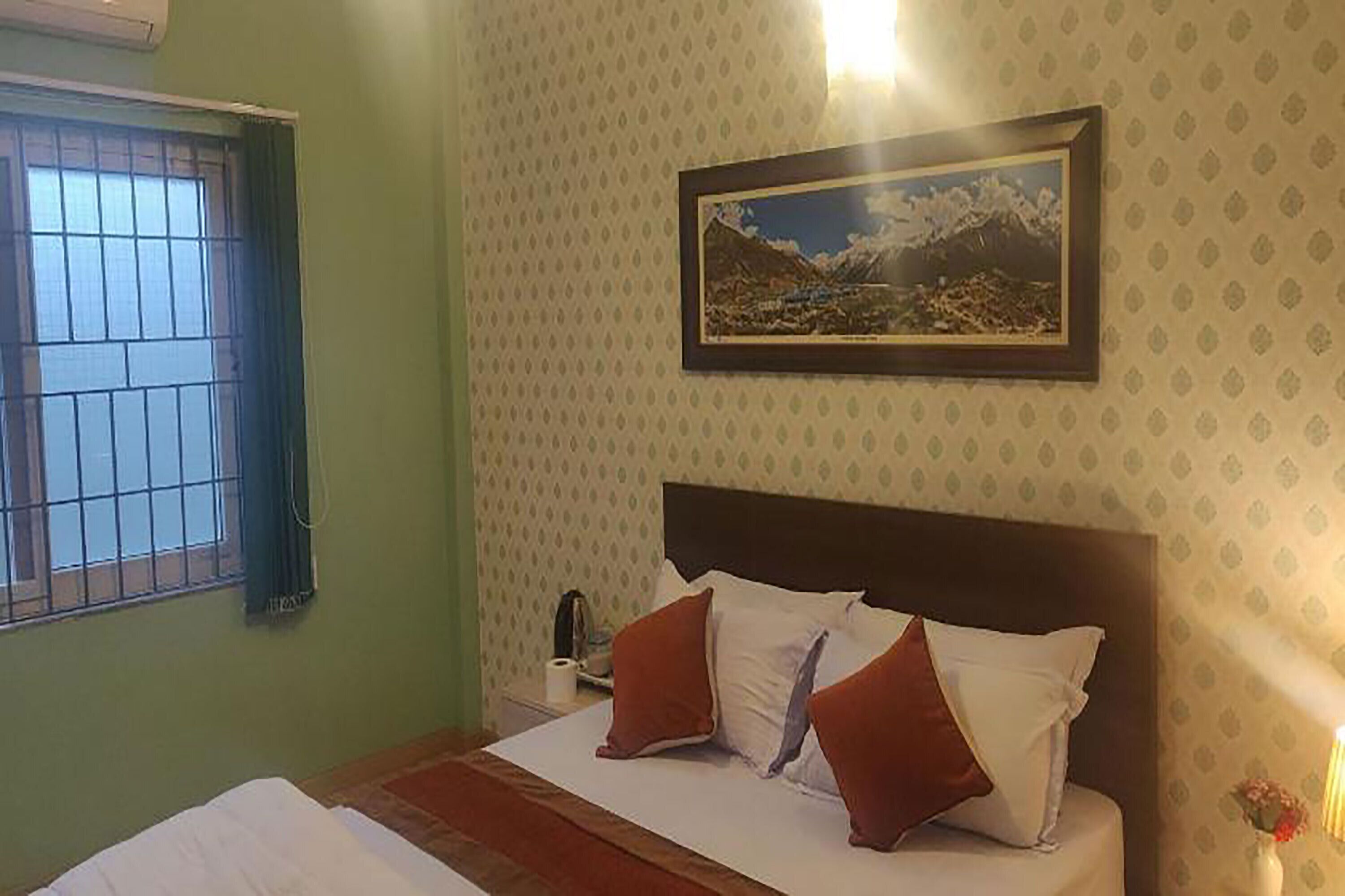 Ein Hotelzimmer mit Bett, einem Fenster mit Gitterstäben, einem gerahmten Bild an der Wand und einem Nachttisch mit Lampe.