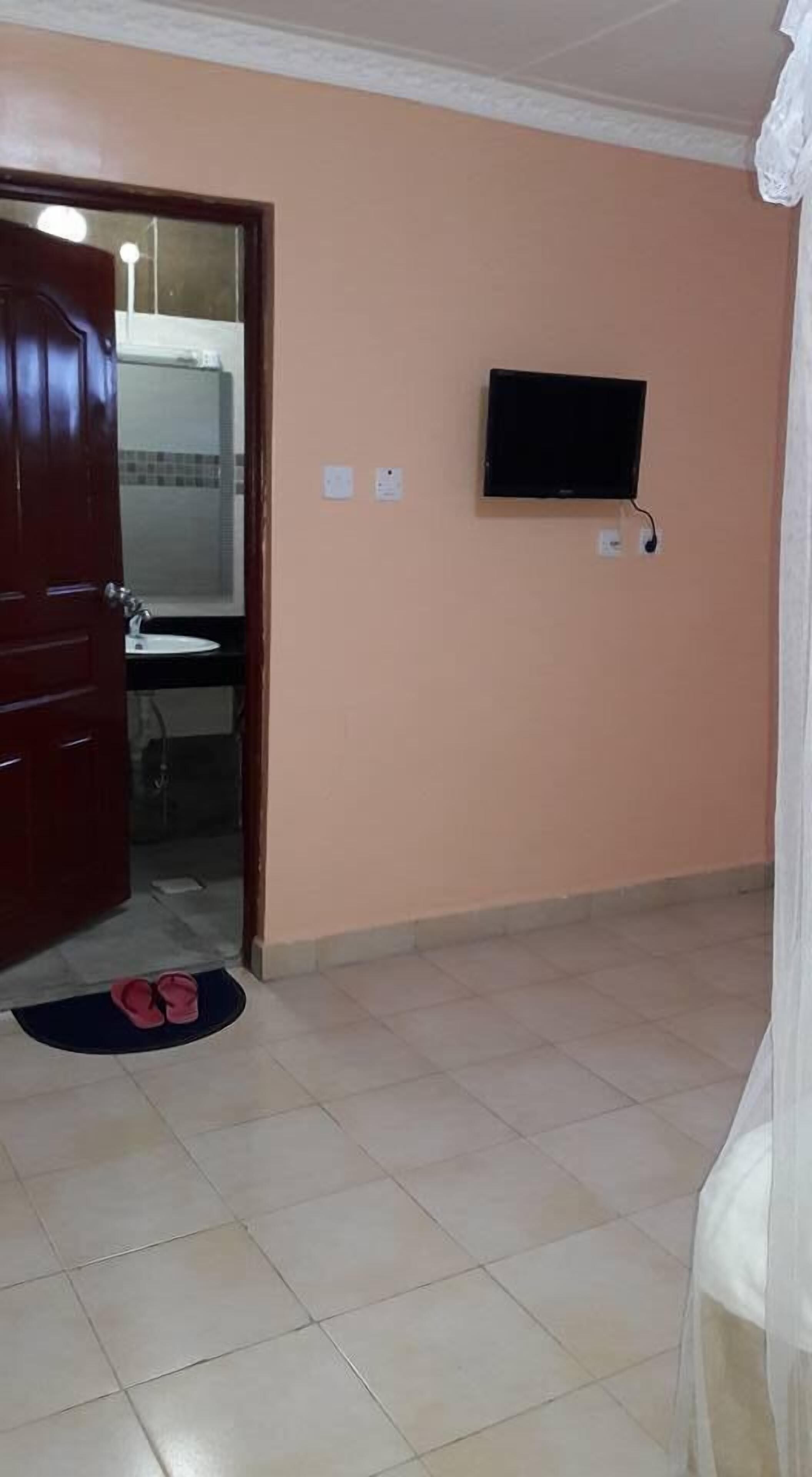 Ein Zimmer mit Holztür, Fliesenboden, einer kleinen Matte, einem an der Wand montierten Fernseher und einem durch die Tür sichtbaren Badezimmer.