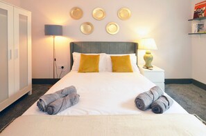 A neatly made bed with white and yellow pillows, a grey headboard, and two rolled towels on the bed. - Townhouse Plus @ London Road Stoke (Stoke-on-Trent)