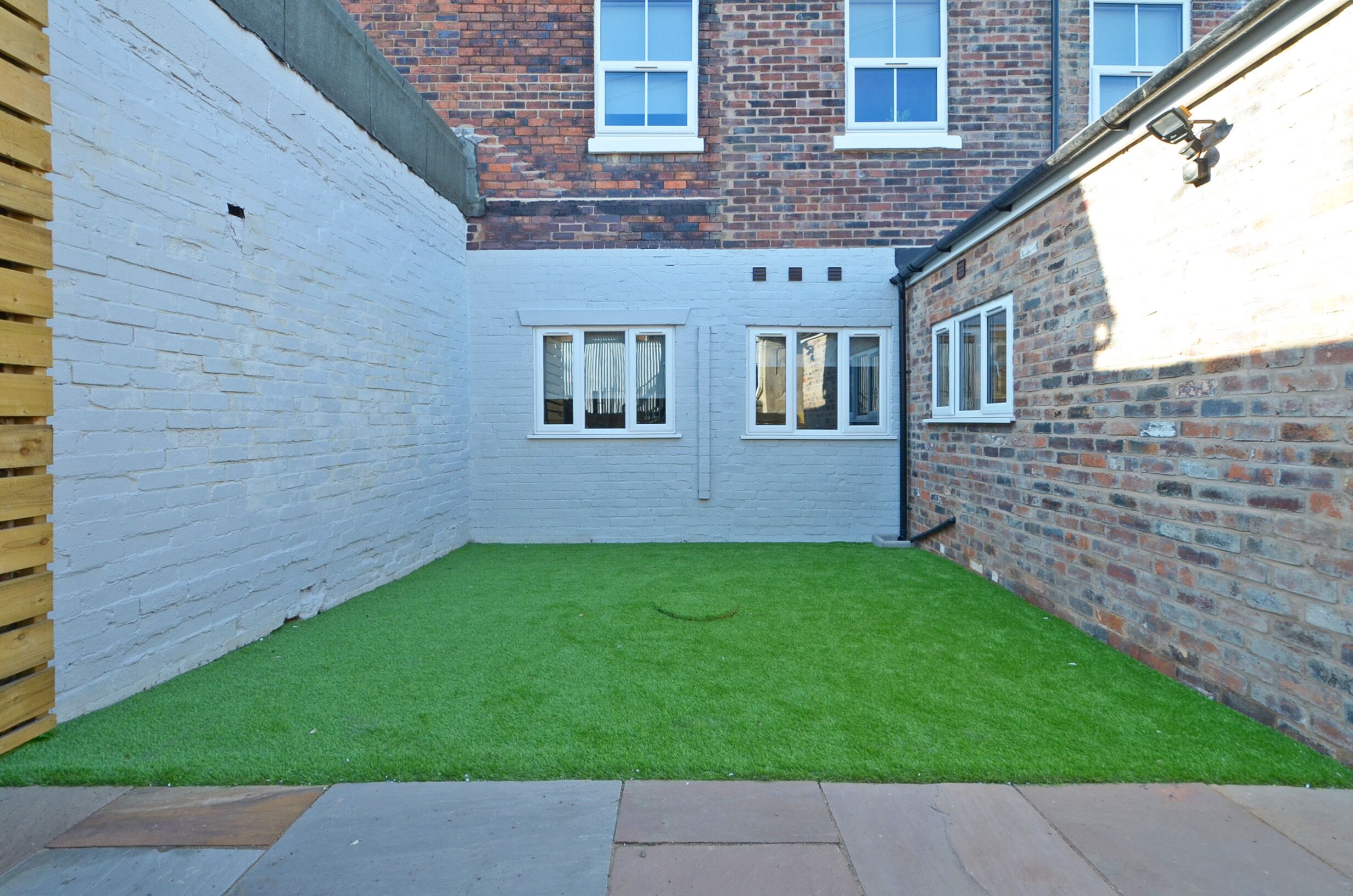 A small, enclosed courtyard with a green lawn, white brick walls, and a wooden fence.