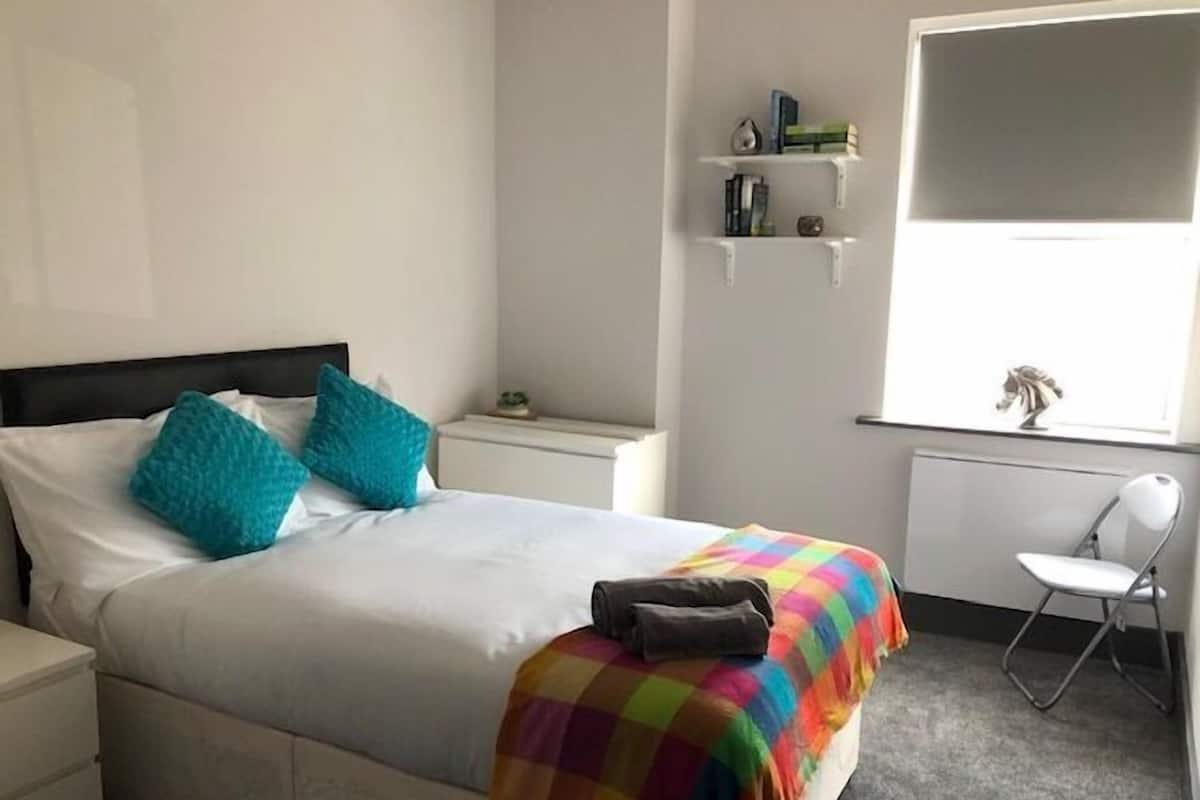 A neatly made bed with a colorful blanket, a white chair, and a small shelf with books and a decorative item.