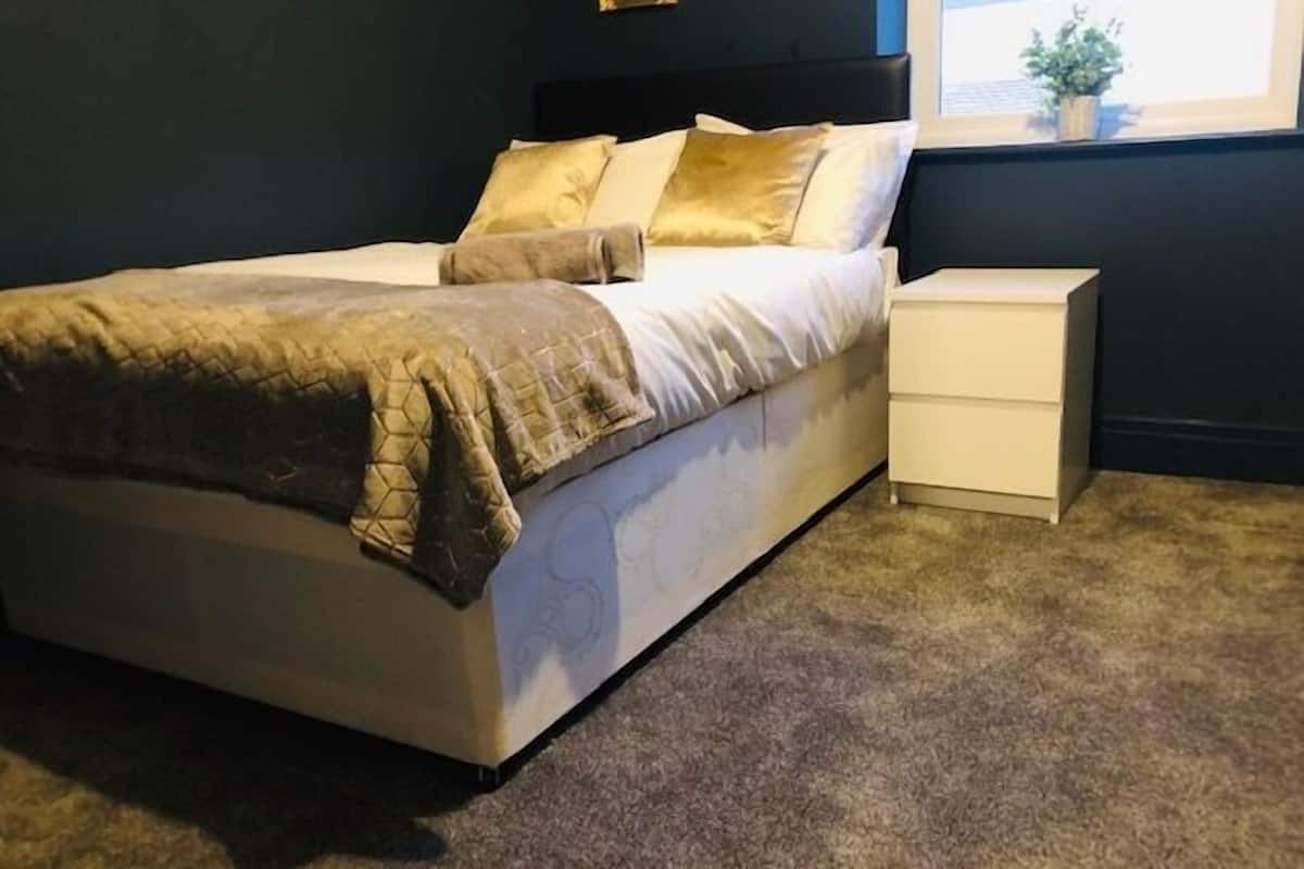 A neatly made bed with a quilted cover, a bedside table with drawers, and a window with a view of the outside.