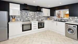 Une cuisine moderne avec des armoires blanches, un dosseret orné d’un motif en chevrons, un four encastré, un lave-vaisselle et une fenêtre donnant sur un bâtiment en briques.