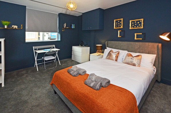 A bedroom with a bed, a desk, and a chair. There are towels on the bed and a lamp on the nightstand.