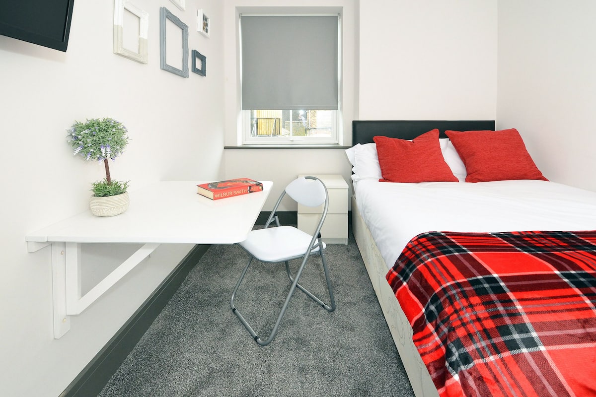 A bedroom with a bed, a desk, a chair, a small potted plant, and a window with a blind.