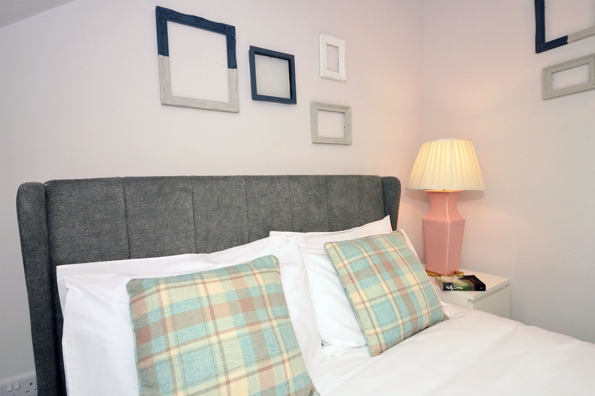 A neatly made bed with white linens and plaid pillows, a grey upholstered headboard, and a bedside table with a lamp and a book.