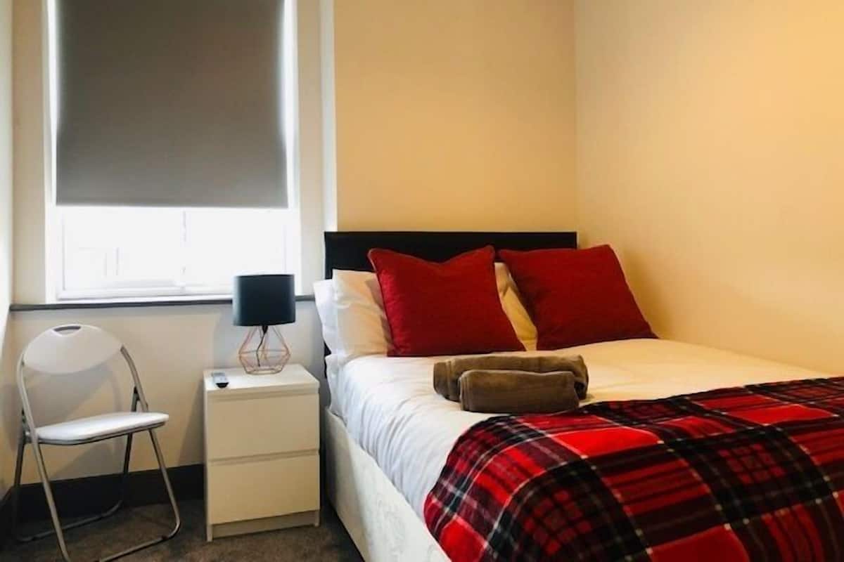 A bedroom with a bed, a nightstand, a chair, a window with blinds, and a lamp.