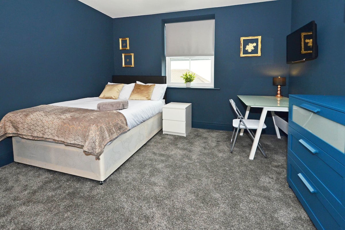 A bedroom with a bed, bedside table, desk, chair, dresser, and window with blinds.