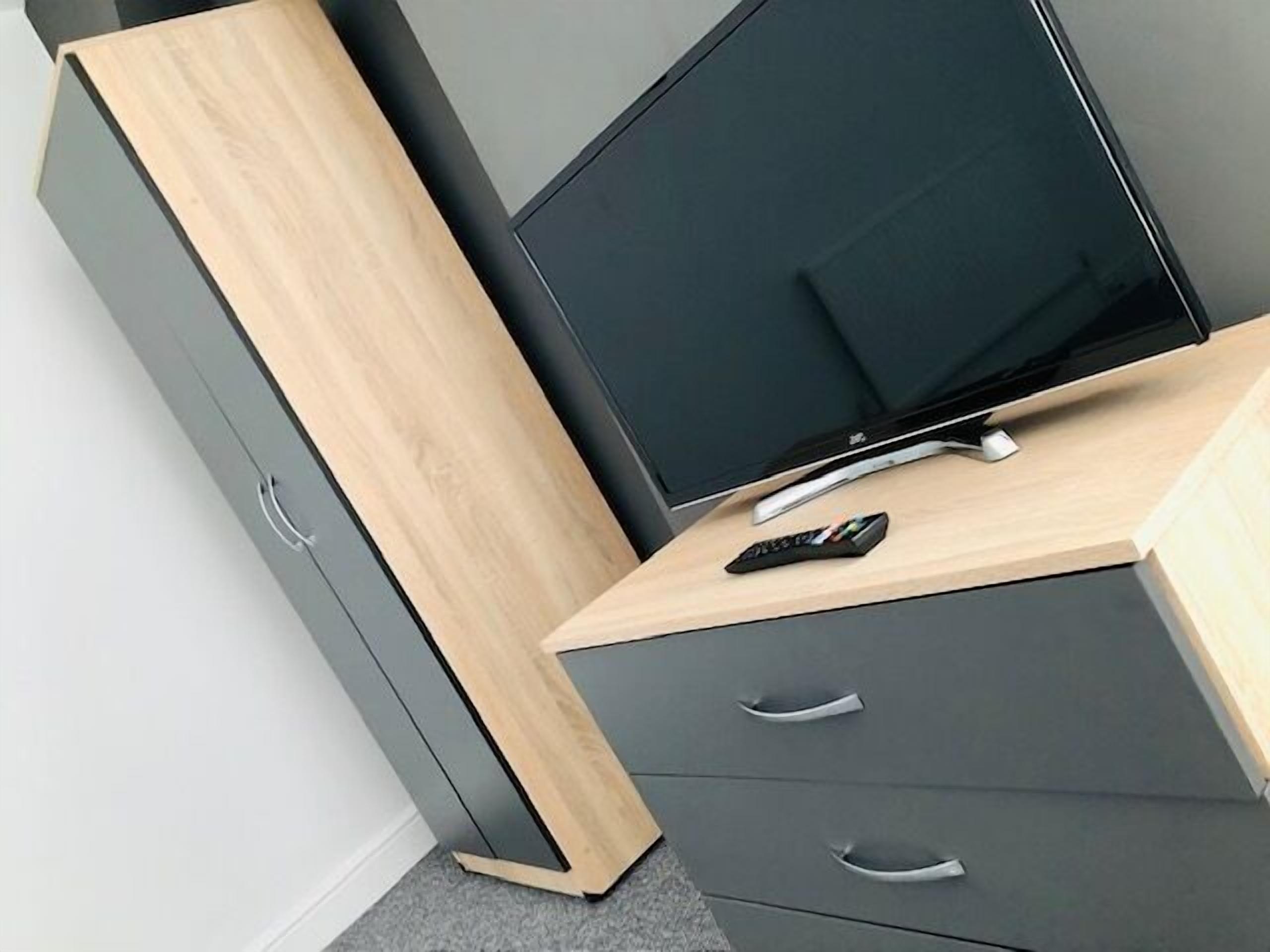Une chambre moderne avec un téléviseur à écran plat posé sur un bureau en bois, une commode grise avec des tiroirs et une télécommande sur le bureau.