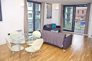A modern living room with a glass dining table, white chairs, a grey sofa with blue cushions, and large sliding glass doors leading to a balcony. - Manchester Ancoats (Manchester)