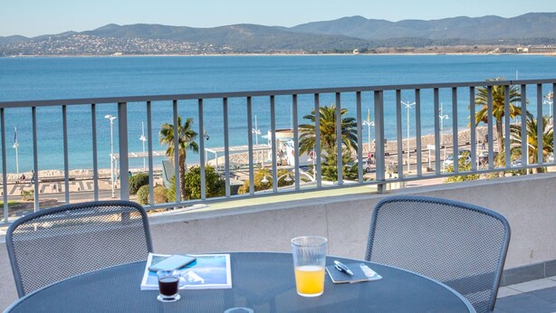 A balcony with a table, chairs, and a drink. The view includes a beach and mountains.