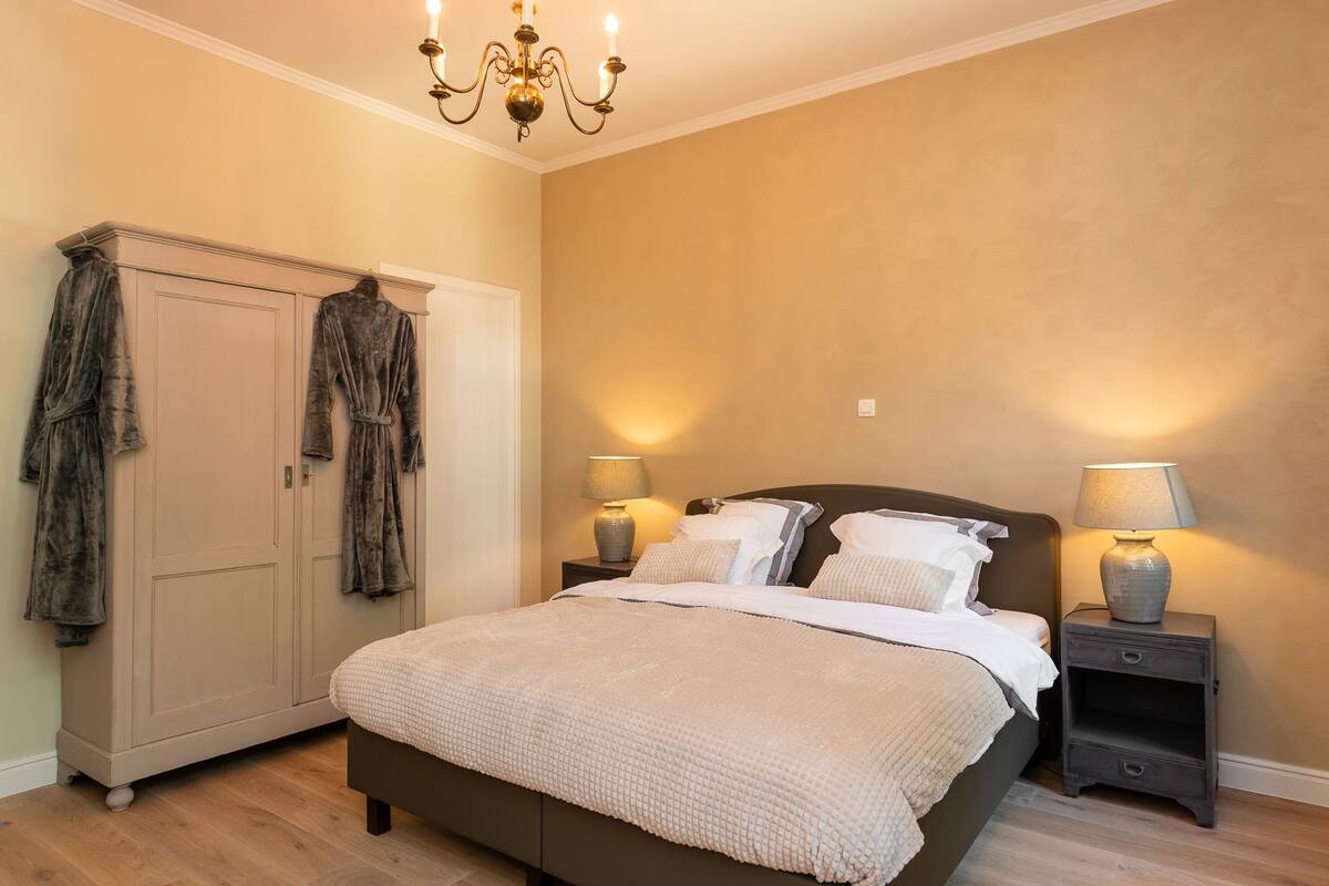 A bedroom with a large bed, two bedside tables with lamps, a wardrobe, and a chandelier.