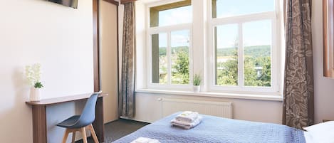 A hotel room with a bed, a desk, a chair, and a window with a view of greenery.