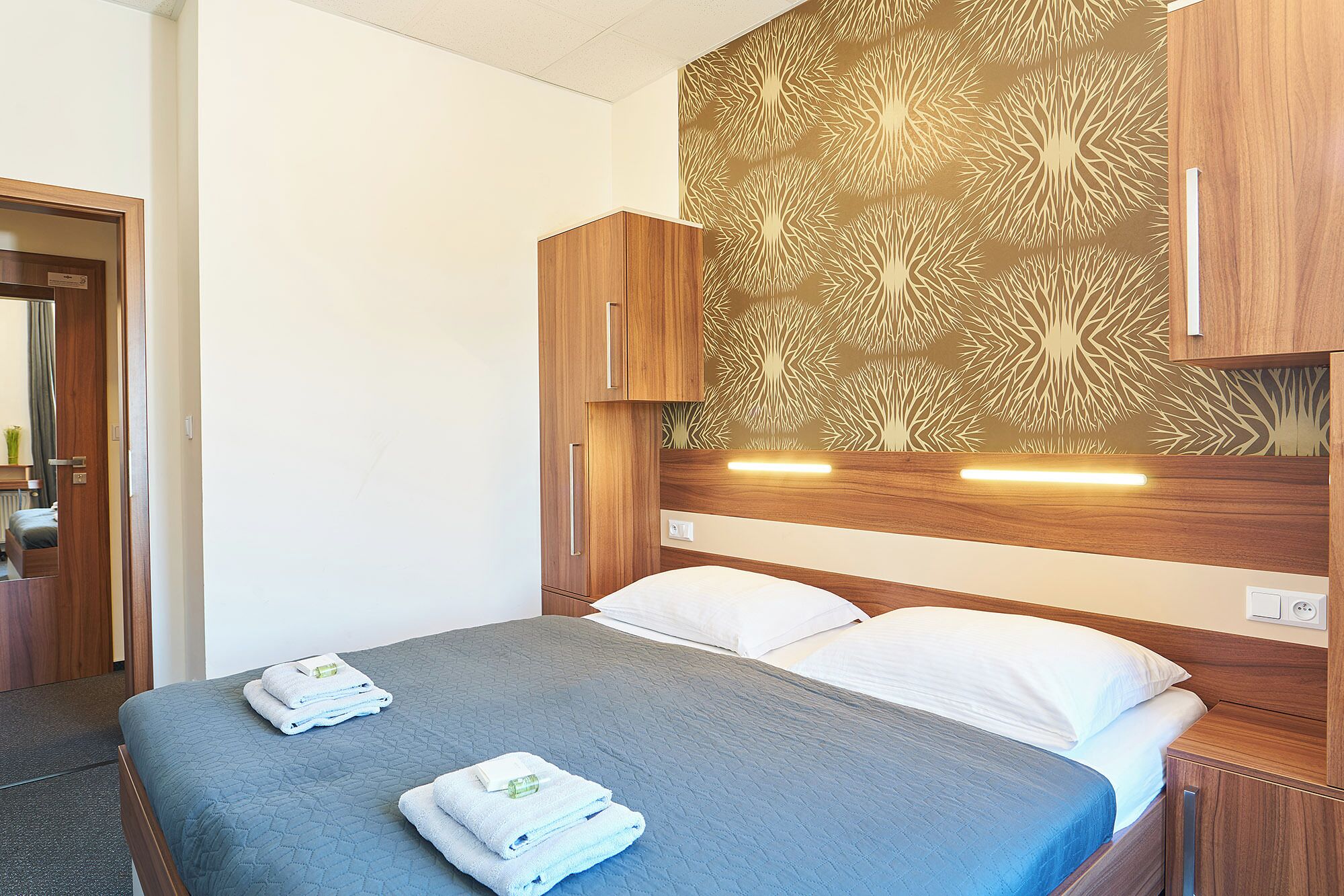 A hotel room with a bed, wooden headboard, and a wall with a tree pattern.