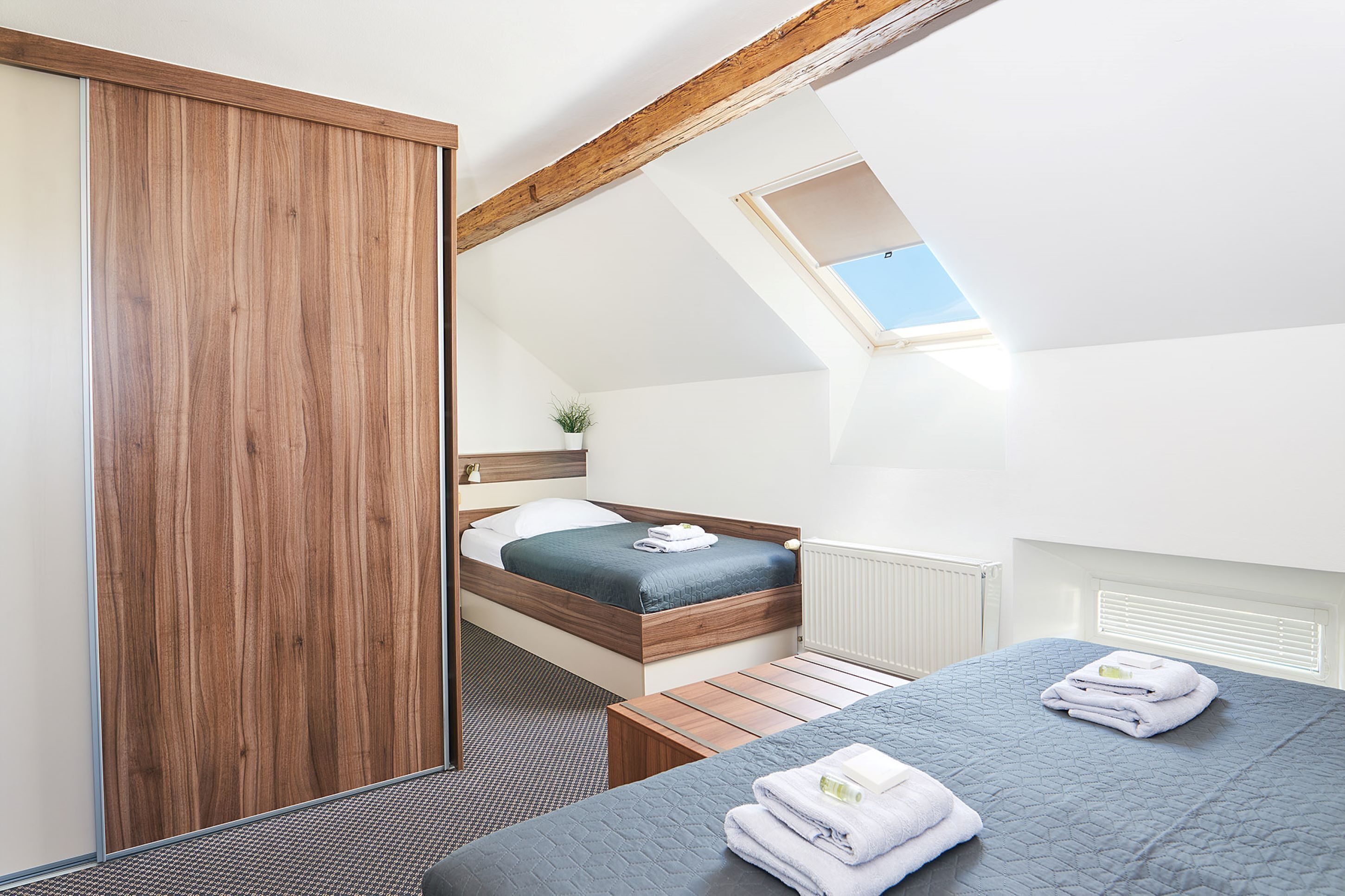 A modern bedroom with a sloped ceiling, a skylight, a wooden wardrobe, a bed with a blue mattress, and a bedside table with towels.