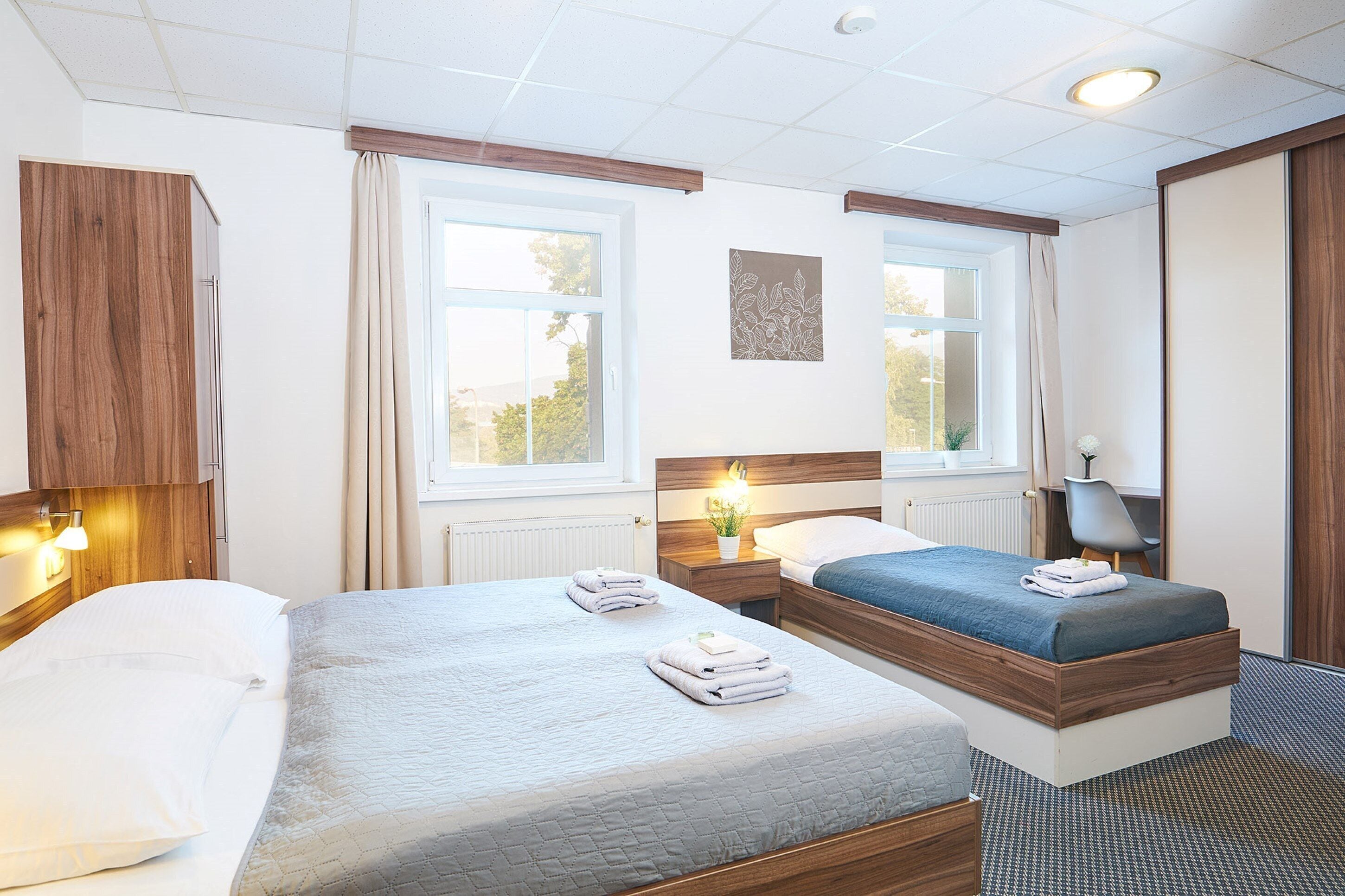 A hotel room with two beds, a wooden wardrobe, a desk with a chair, and a window with curtains.