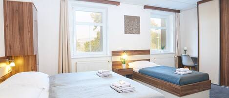 A hotel room with two beds, a wooden wardrobe, a desk with a chair, and a window with curtains.