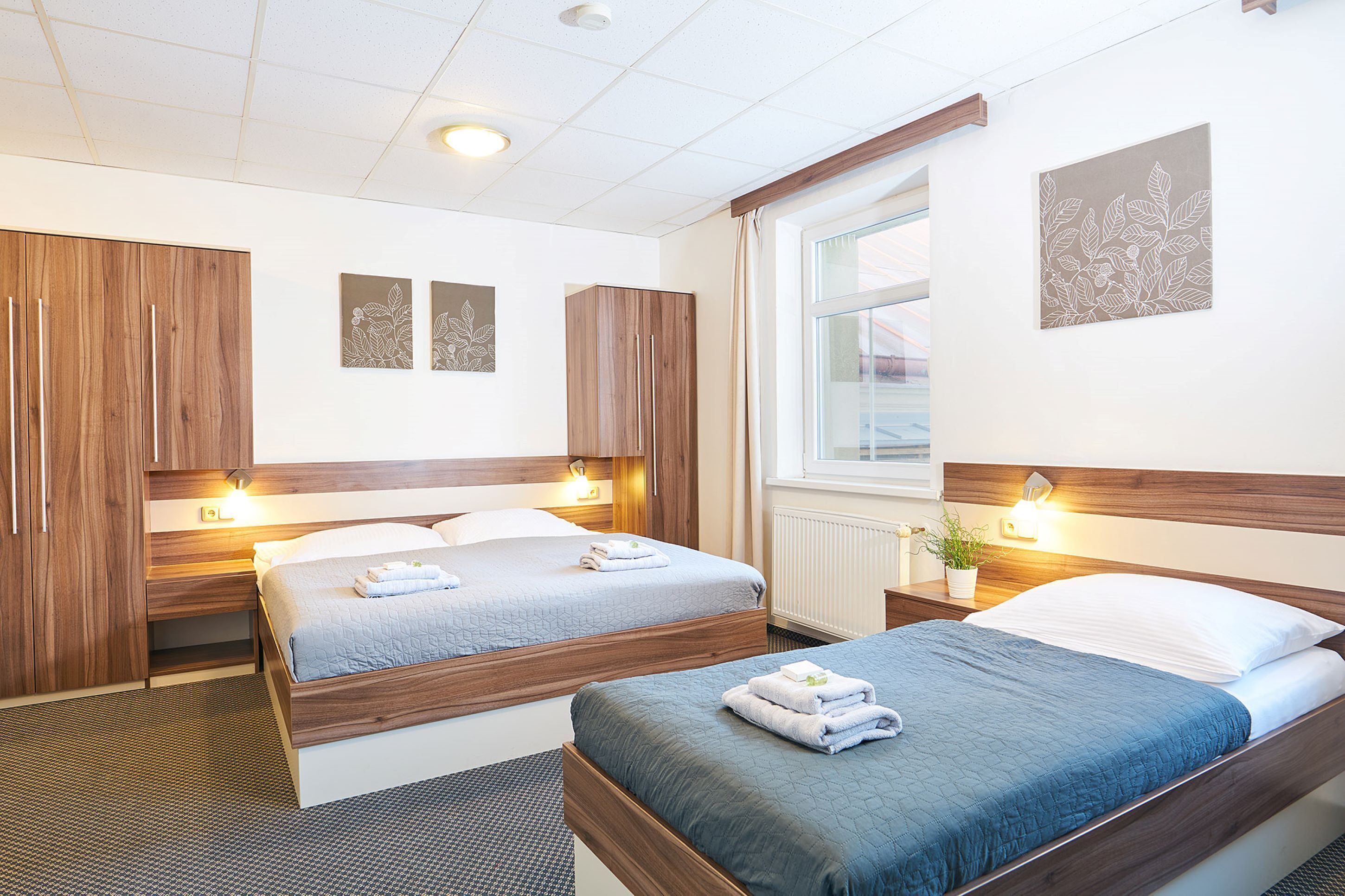 A hotel room with two beds, wooden headboards, bedside tables with lamps, a window with curtains, and wall art.
