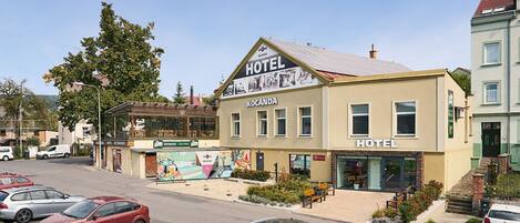 A two-story hotel with a sign reading "HOTEL KOCANDA" and a restaurant below. There are parked cars in front and a pedestrian walkway with benches.