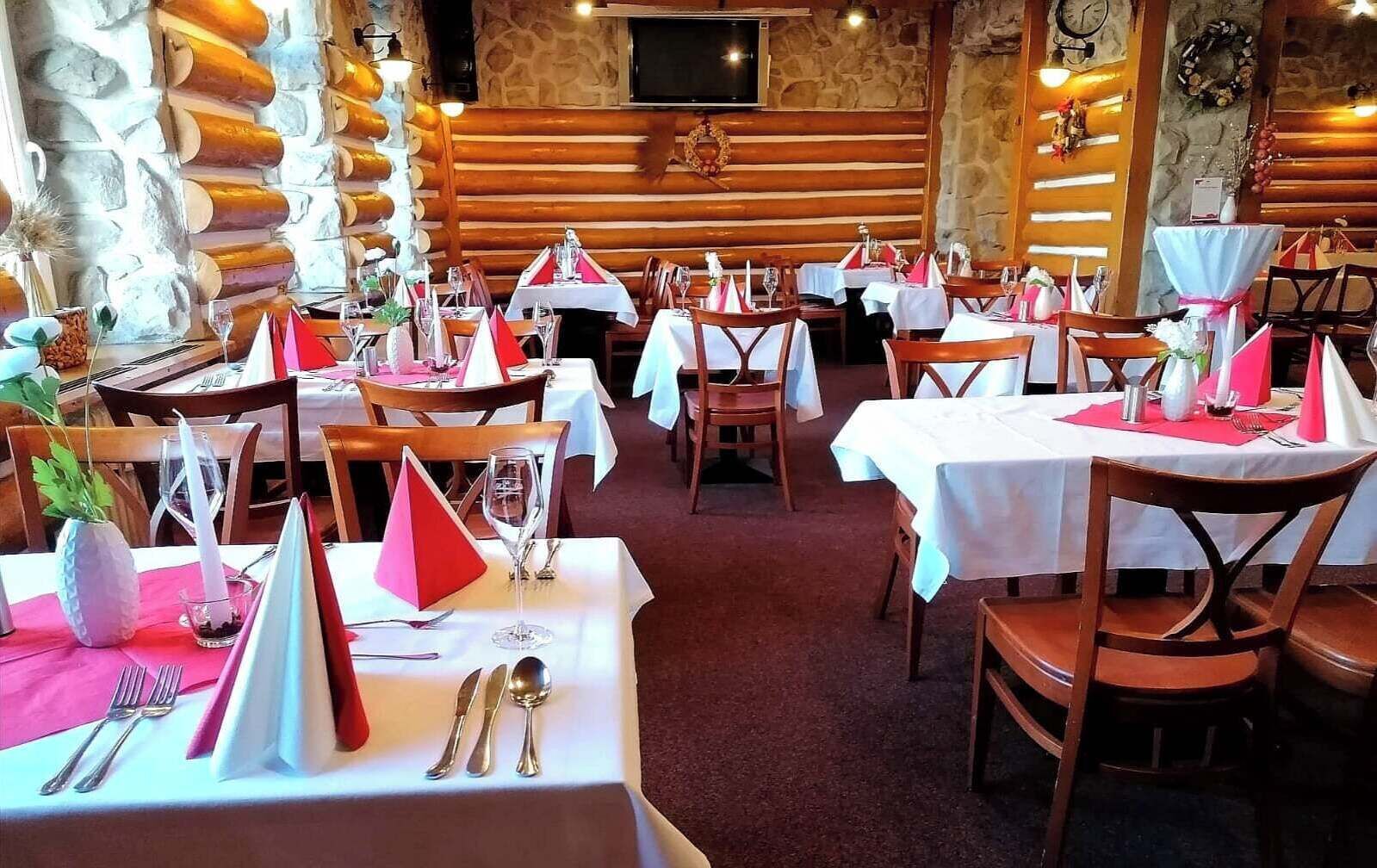 Un restaurante con paredes de madera y detalles de piedra, mesas dispuestas con manteles blancos y servilletas rojas, y un reloj en la pared.