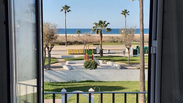 Blick von einem Balkon mit Metallgeländer auf eine angelegte Grünfläche mit Palmen, einem Weg und einem Strand im Hintergrund.