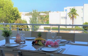 Une table à manger dressée avec des assiettes, des couverts, une bouteille et un panier de fruits, sur un balcon offrant une vue sur des immeubles et des arbres.