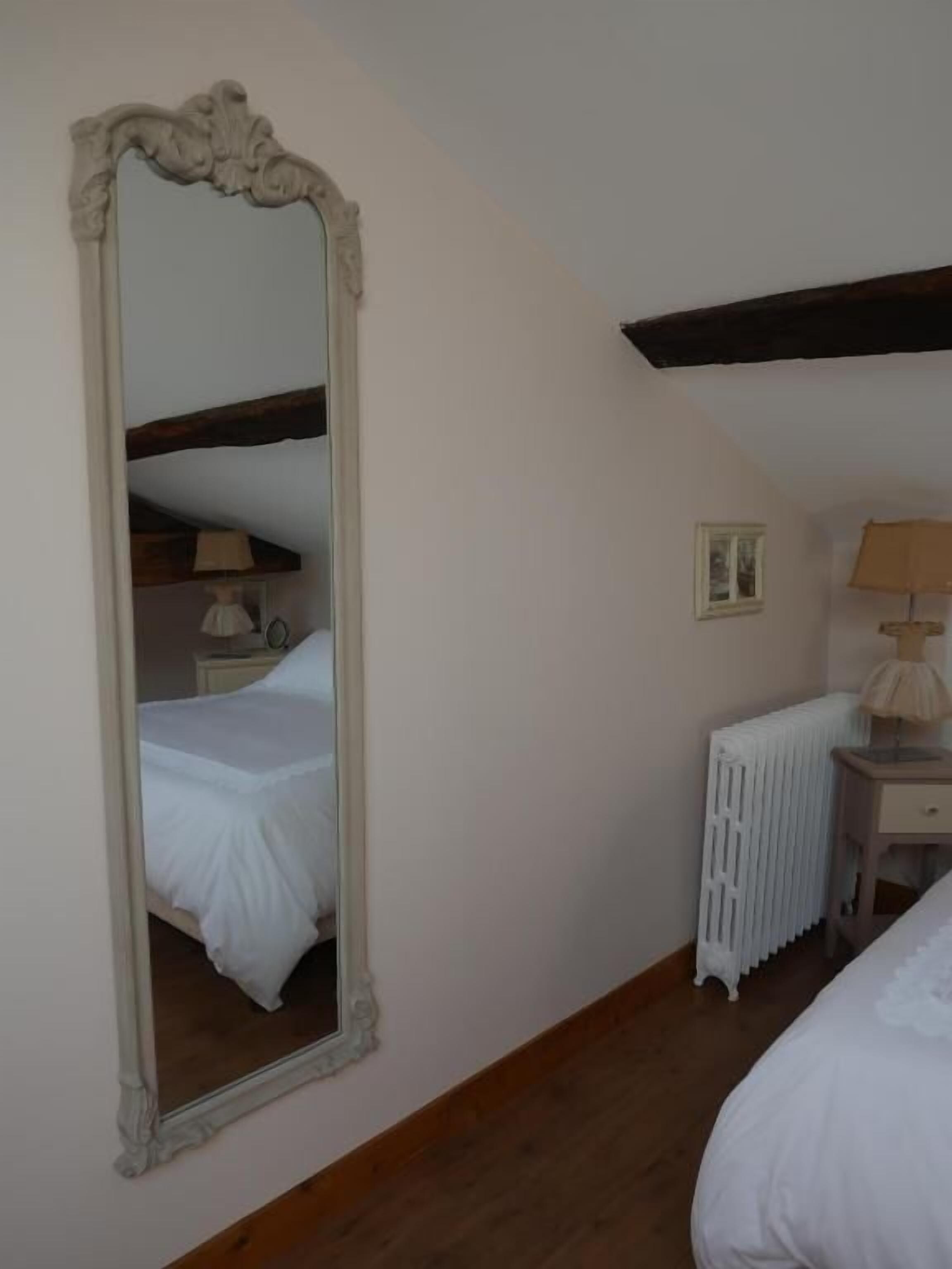 A bedroom with a large mirror, a bed, a nightstand, a lamp, and a radiator.