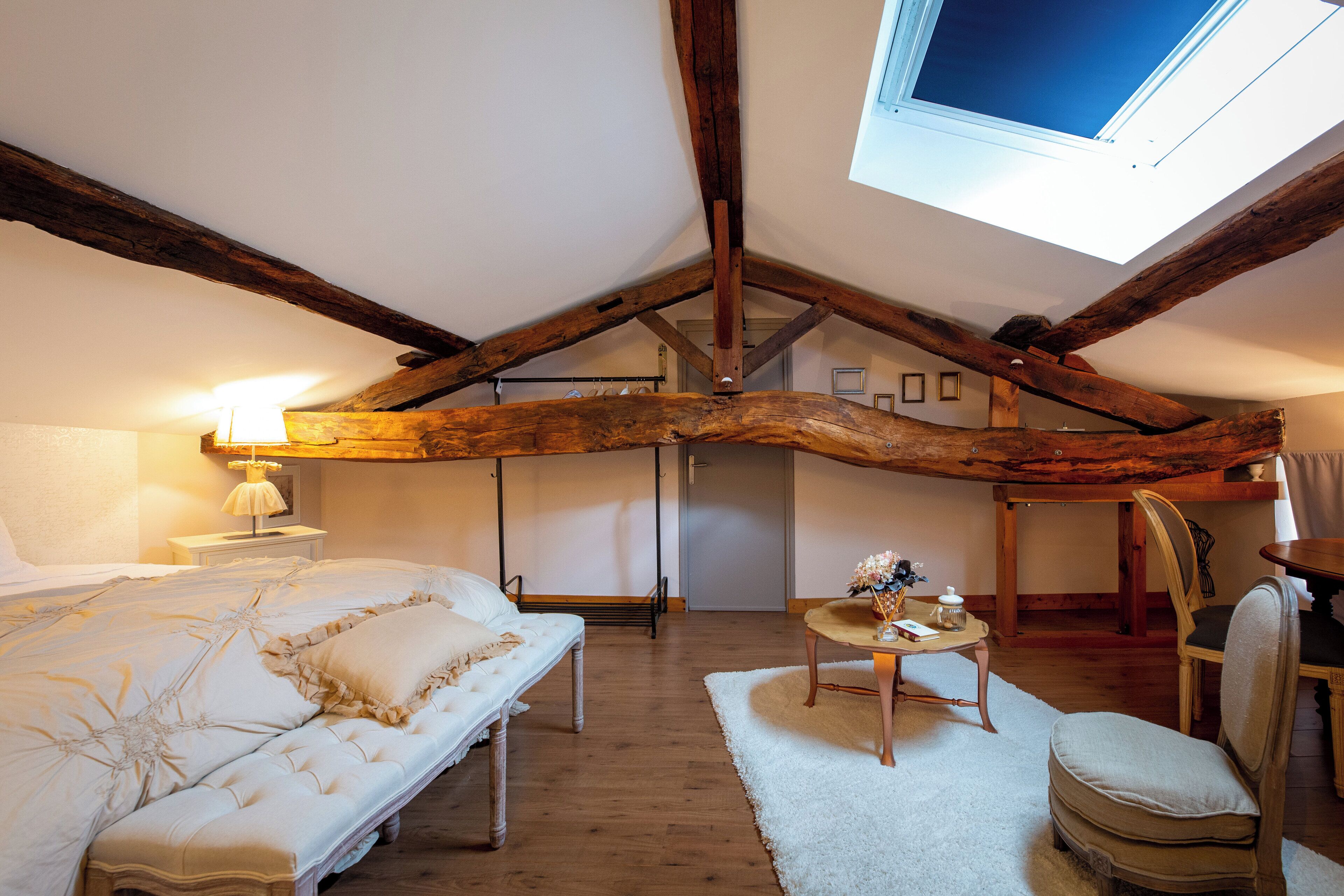 A bedroom with a sloped ceiling, exposed wooden beams, a bed with white bedding, a small table with a lamp, and a chair.