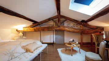 A bedroom with a sloped ceiling, exposed wooden beams, a bed with white bedding, a small table with a lamp, and a chair.