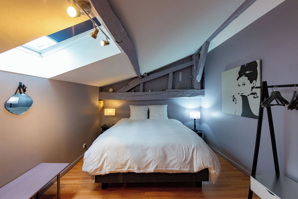 Een slaapkamer met dakraam, een bed met witte beddengoed, nachtlampen, een bankje en een kapstok aan de muur.