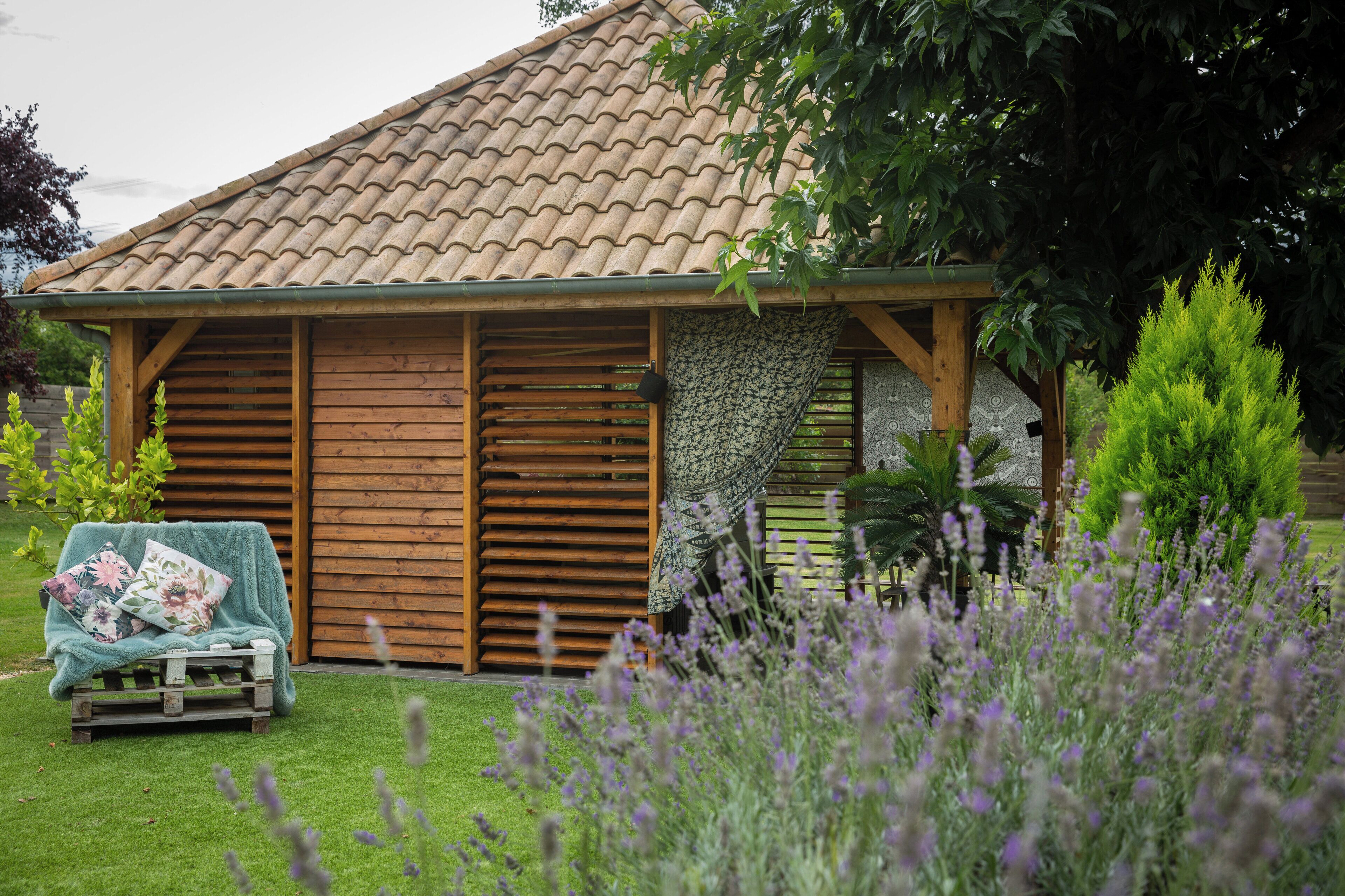 Een houten hutje met pannendak, een overdekte zithoek met kussens, en een tuin vol lavendel.